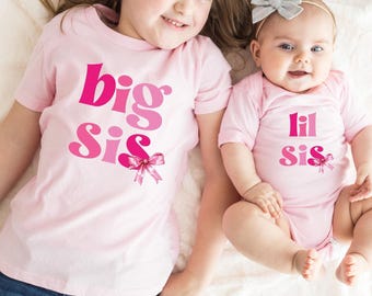 May include: Two children wearing matching pink shirts and onesie. The shirt says "big sis" and the onesie says "lil sis" in pink lettering. Both outfits have a pink bow graphic. The children are smiling.