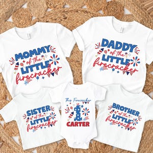 May include: White t-shirts and a onesie with patriotic designs. The shirts read "Mommy," "Daddy," "Sister," and "Brother" with "of the Little Firecracker" in red and blue. A onesie says "This Firecracker is 1" and "Carter."
