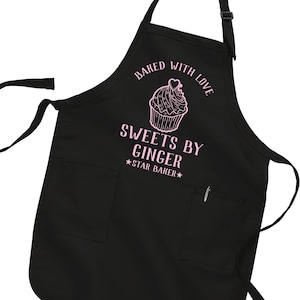 May include: Black apron with pink text and a cupcake graphic. The text reads "Baked with Love" above a cupcake illustration, and "Sweets by Ginger" with "★Star Baker★" below. The apron has two front pockets and adjustable straps.