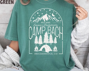 May include: Light green t-shirt with a white graphic. The graphic includes mountains, trees, a tent, and the text "Emma's Bachelorette Camp Bach July 2025 Breckenridge, CO."