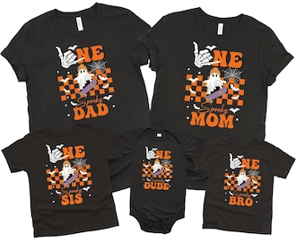 May include: Black Halloween family t-shirts and a onesie. Each shirt features a skeleton hand, a ghost on a skateboard, and the words "ONE" and "Spooky" with family member titles: Dad, Mom, Sis, Dude, and Bro. Orange and black checkered background.