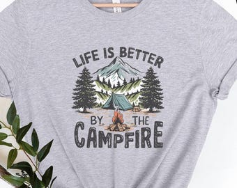 May include: A heather gray t-shirt with a camping graphic. The graphic features a mountain scene with a tent, trees, and a campfire, with the text "LIFE IS BETTER BY THE CAMPFIRE" in a dark font.