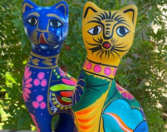 May include: Two ceramic cat figurines, one blue with pink and yellow details, the other yellow with green, orange, and blue details.