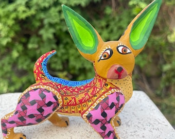 May include: A colorful wooden carved chihuahua figurine with green ears, pink and black patterned legs, and a yellow, red, and blue patterned body.