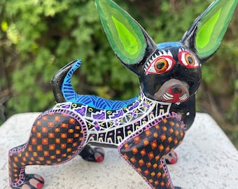 May include: A hand-painted wooden figurine of a chihuahua dog. The dog is black with bright green ears, orange eyes, and a pink nose. The body is decorated with colorful geometric patterns in black, orange, pink, blue, and purple.