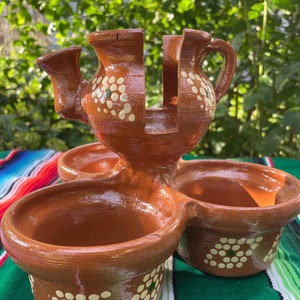 May include: A terracotta serving dish with three bowls and a central teapot-shaped container. The bowls and teapot are decorated with white dots and green accents.