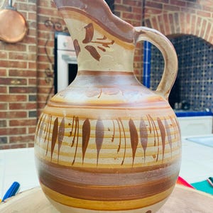 May include: A handcrafted, ceramic pitcher with a rounded base and a pouring spout. The pitcher features a beige base with brown and tan horizontal stripes and decorative leaf-like patterns. The handle is curved, and the pitcher is set on a wooden surface.