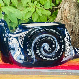 May include: A ceramic snail-shaped planter in glossy dark blue with white and light blue decorative patterns. The planter has a wide opening for plants and sits on a colorful striped surface. The snail's eyes are on stalks.
