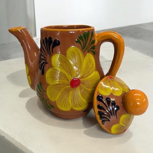 May include: A terracotta teapot with a floral design in shades of yellow, red, green, and black. The teapot has a lid with a knob on top.