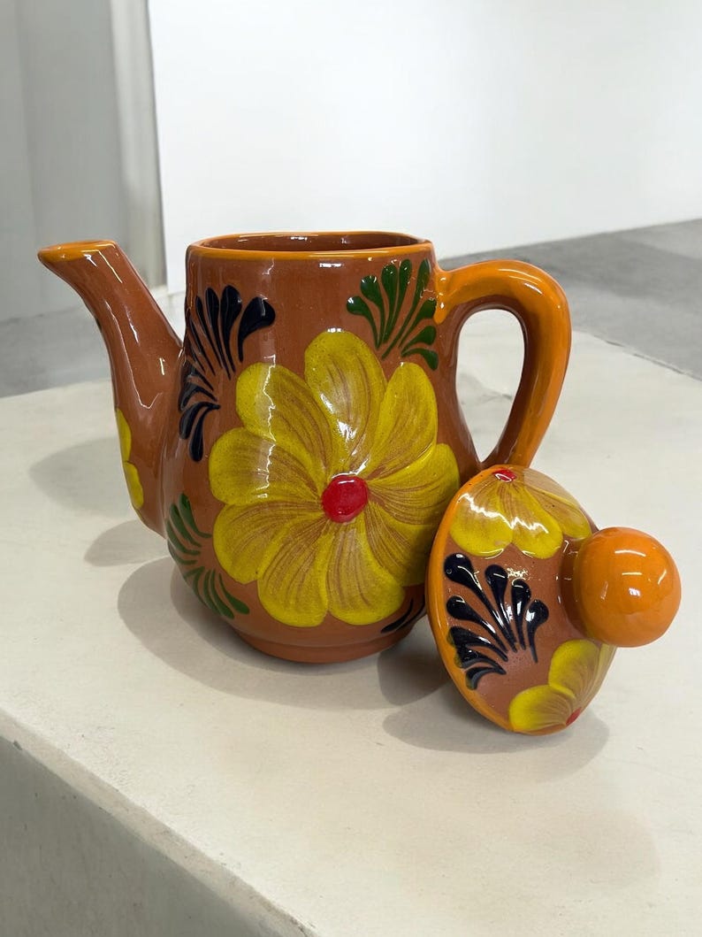 May include: A terracotta teapot with a floral design in shades of yellow, red, green, and black. The teapot has a lid with a knob on top.