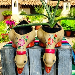 May include: Two hand-painted, beige ceramic planters shaped like geese, each with a red collar and beak. The planters are decorated with floral designs in pink, red, and green. Aloe vera plants are planted in the planters.