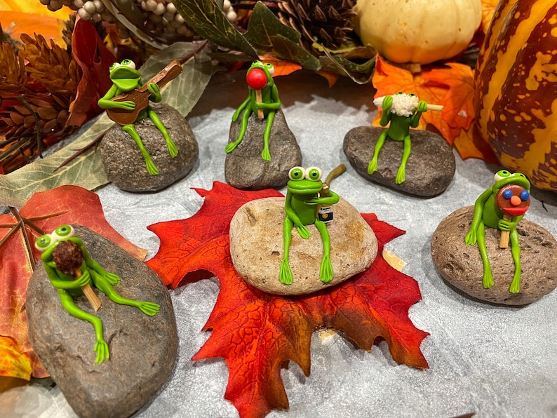 May include: Six green frog figurines, each sitting on a gray rock, with various accessories. The frogs are holding a guitar, a lollipop, a popsicle, a bottle, a corn cob, and a stick. The rocks are surrounded by fall leaves.