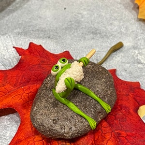 May include: A green frog figurine with large eyes sits on a gray rock, holding a white stick with a white fluffy substance on the end. The rock is on a red and orange leaf.