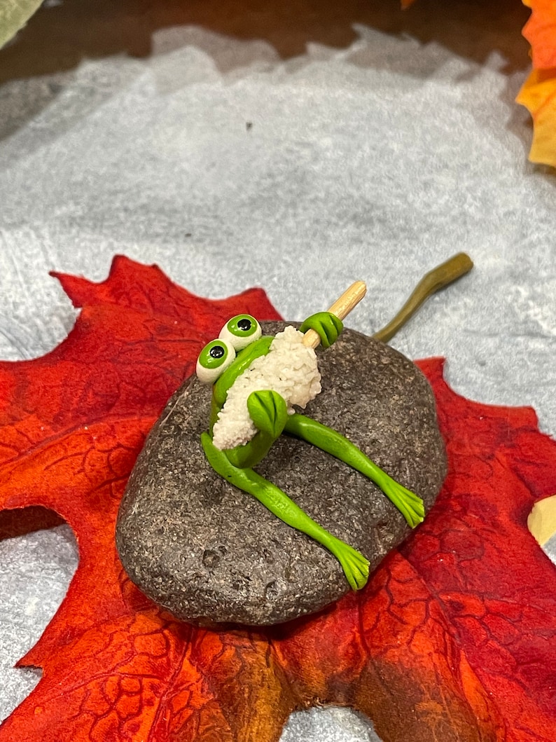 May include: A green frog figurine with large eyes sits on a gray rock, holding a white stick with a white fluffy substance on the end. The rock is on a red and orange leaf.
