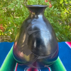 May include: A black ceramic vase with a rounded body and a narrow neck. The vase features etched geometric designs. The background includes green foliage and a colorful striped textile.