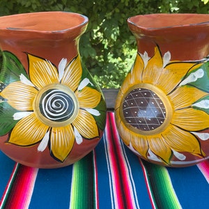 May include: Two terracotta pottery vases with sunflower designs. The vases are painted with yellow, white, green, and brown accents. The vases are sitting on a colorful striped blanket.