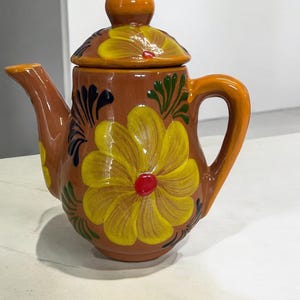 May include: A small brown ceramic teapot with a lid and a handle. The teapot is decorated with yellow flowers and green leaves.