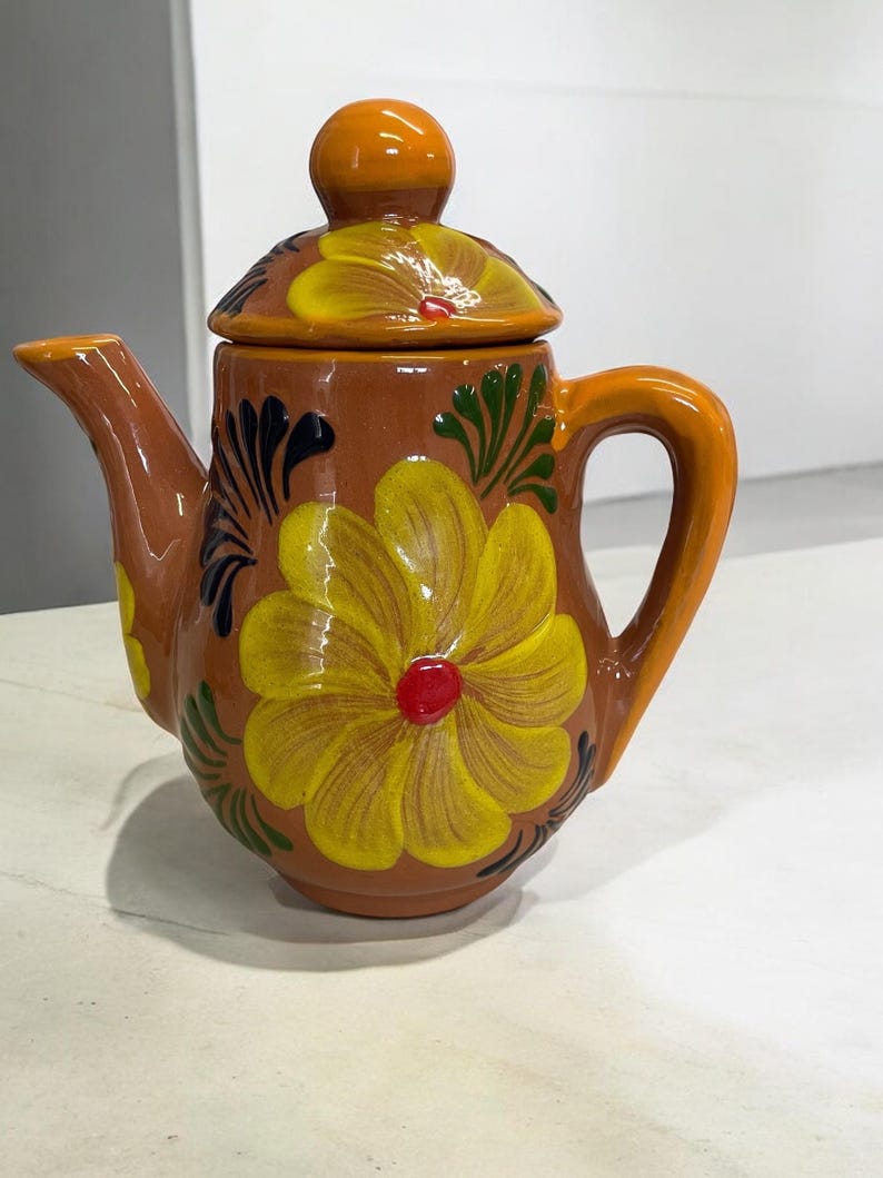 May include: A small brown ceramic teapot with a lid and a handle. The teapot is decorated with yellow flowers and green leaves.