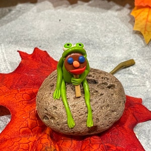 May include: A green frog figurine with blue eyes and a red mouth sits on a gray rock holding a brown stick with a red, white, and blue lollipop on top. The frog is sitting on a red and orange leaf.