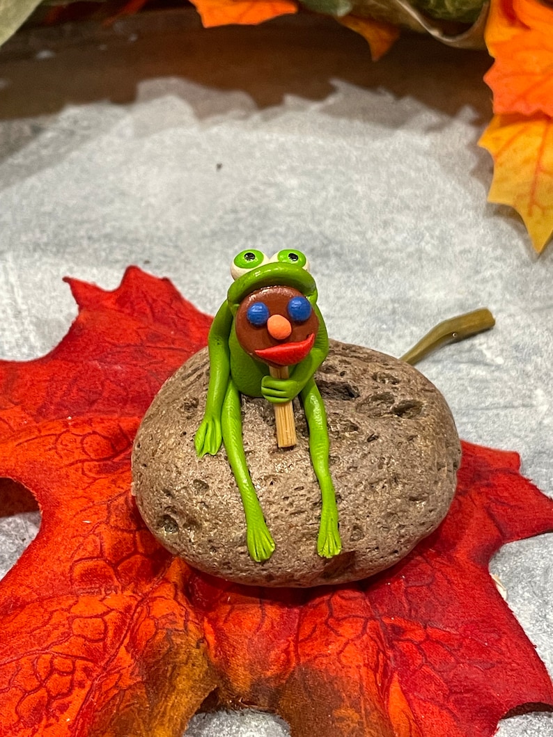 May include: A green frog figurine with blue eyes and a red mouth sits on a gray rock holding a brown stick with a red, white, and blue lollipop on top. The frog is sitting on a red and orange leaf.