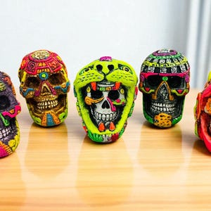May include: Five colorful Day of the Dead sugar skulls with intricate designs. The skulls are painted in vibrant hues of pink, green, yellow, and orange, with detailed patterns and facial features. They are arranged on a light brown wooden surface.