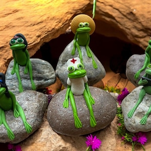 May include: A collection of whimsical green frog figurines, each posed on a gray stone. Some frogs wear hats, a nurse's cap, or hold a tablet. The background features a natural rock formation and small purple flowers.