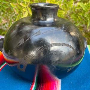 May include: Black ceramic vase with a round body and a narrow neck. The vase has a geometric design carved into the surface.