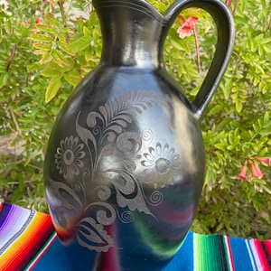 May include: Black ceramic pitcher with a floral design carved into the surface. The pitcher has a wide mouth and a handle.