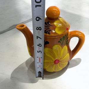May include: A small, brown ceramic teapot with a yellow floral design. The teapot has a round lid with a knob on top. The teapot is 7 inches tall.