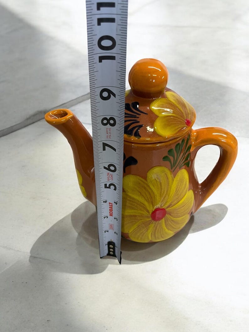 May include: A small, brown ceramic teapot with a yellow floral design. The teapot has a round lid with a knob on top. The teapot is 7 inches tall.