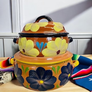 May include: Two stacked terracotta casserole dishes with floral designs. The top dish has yellow flowers and the bottom dish has blue flowers.