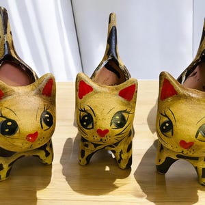 May include: Three ceramic cat planters. Each planter is painted with a tan base color, black spots, and red accents on the ears and nose. The cat's eyes are large and black with white highlights. The tail of each cat is raised.