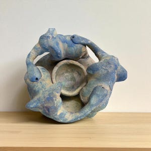 May include: A ceramic sculpture featuring three figures in a circular arrangement, each with a blue and beige mottled finish. The figures appear to be embracing a central bowl, creating a unique artistic composition. The sculpture is displayed on a wooden surface.