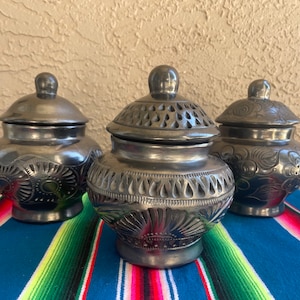 May include: Three black ceramic jars with lids, each with a different carved design. The jars are sitting on a colorful striped blanket.