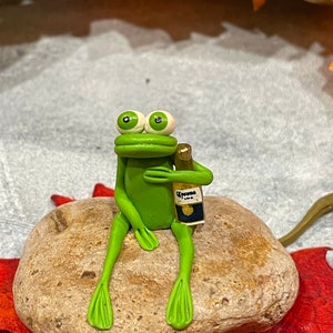 May include: A green frog figurine sitting on a brown rock, holding a small bottle of Corona beer.