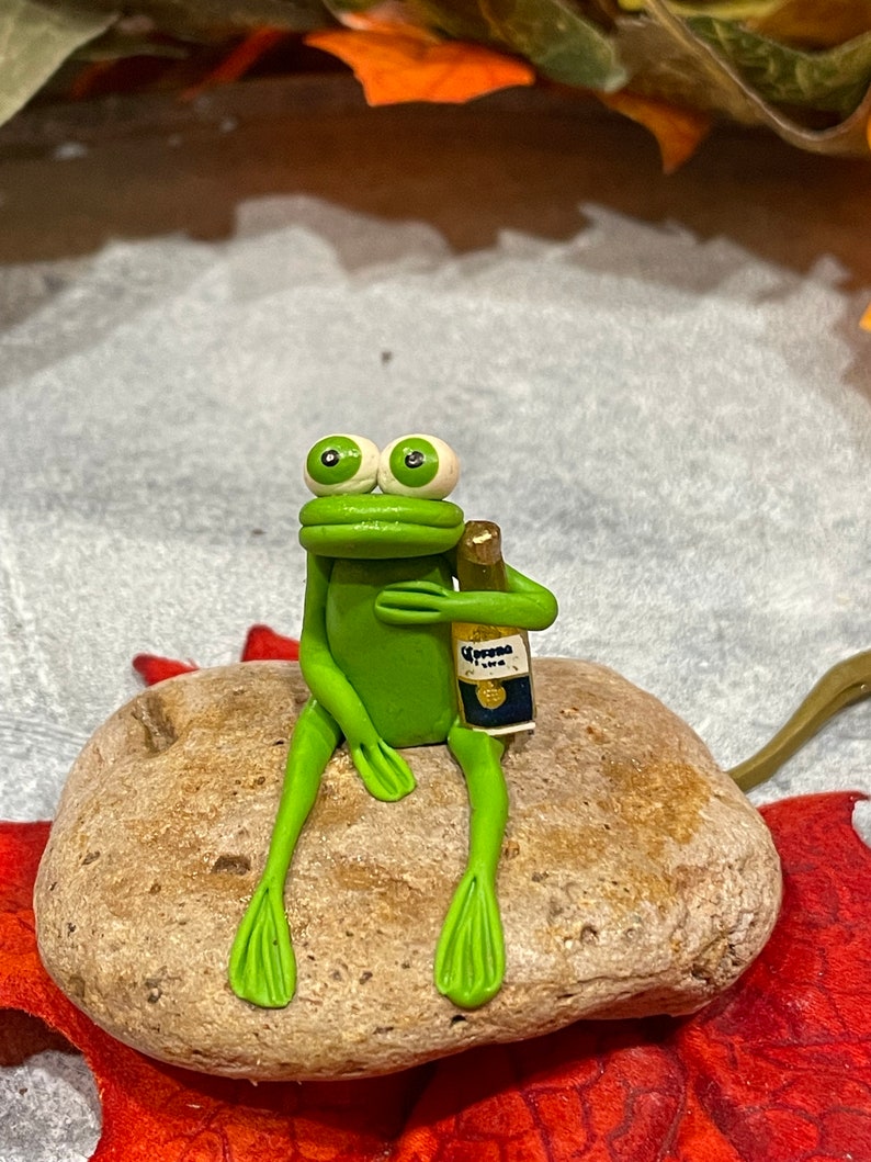 May include: A green frog figurine sitting on a brown rock, holding a small bottle of Corona beer.