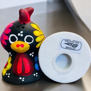 May include: A black ceramic rooster salt and pepper shaker set with colorful painted details. The rooster has a red comb and wattle, yellow eyes, and a black body with pink, yellow, and blue flowers. The shaker has a white base with a "HECHO EN MEXICO" label.