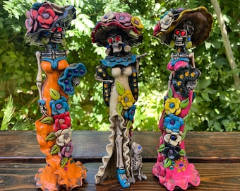 May include: Three colorful Day of the Dead skeleton figurines wearing elaborate hats and dresses with floral and jewel accents. The figurines are standing on a wooden surface.