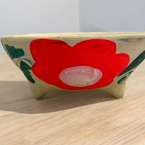 May include: A small, hand-painted ceramic bowl with three legs. The bowl is decorated with a large red flower with a white center and green leaves.