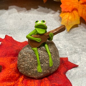 May include: A green frog figurine playing a brown guitar while sitting on a gray rock. The frog has large, white eyes and a wide, open mouth. The rock is surrounded by red and orange fall leaves.