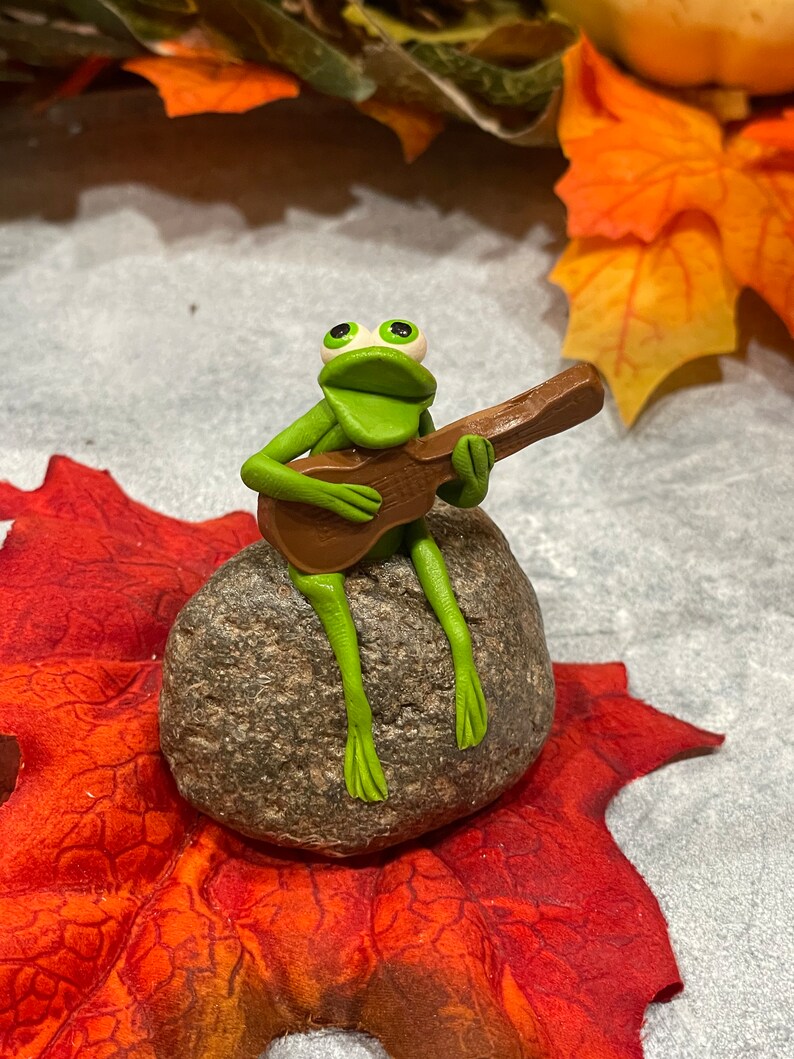May include: A green frog figurine playing a brown guitar while sitting on a gray rock. The frog has large, white eyes and a wide, open mouth. The rock is surrounded by red and orange fall leaves.
