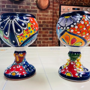 May include: Two hand-painted, colorful ceramic goblets. The goblets feature a dark blue rim and base, with a white stem. The bodies are adorned with intricate floral and geometric patterns in red, orange, yellow, and green. The background includes a brick wall.