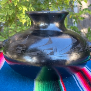 May include: A black ceramic vase with a rounded body and a flared rim. The vase features etched geometric designs around the middle and small handles on either side. The vase is displayed on a blue and striped textile.