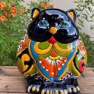 May include: A ceramic cat figurine with a black, blue, green, yellow, orange, and white painted design. The cat is sitting on a wooden surface.