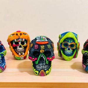 May include: A collection of colorful, decorative skull figurines. Each skull is uniquely painted with vibrant hues of blue, orange, green, and black, featuring intricate patterns and designs. The skulls are displayed on a light brown wooden surface.