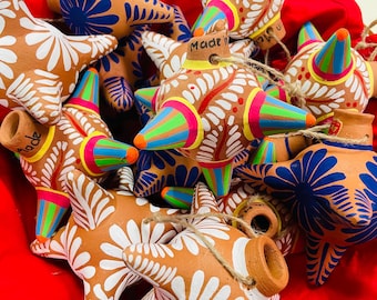 May include: A collection of colorful, handcrafted clay ornaments. The ornaments feature star and pinata shapes, adorned with vibrant paint in shades of blue, orange, pink, green, and white. Each piece has a rustic, artisanal feel, with the word "Made" visible on some.