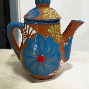May include: A terracotta teapot with a blue lid and floral designs. The teapot is decorated with blue, green, white, and red flowers.