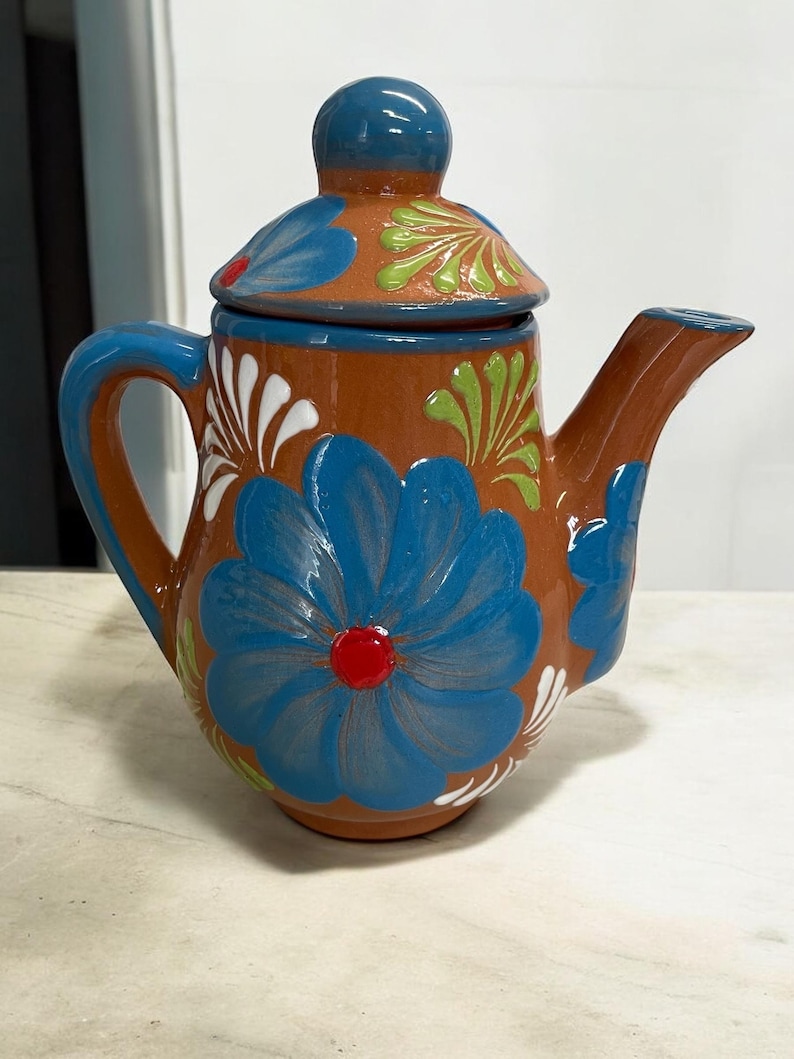 May include: A terracotta teapot with a blue lid and floral designs. The teapot is decorated with blue, green, white, and red flowers.