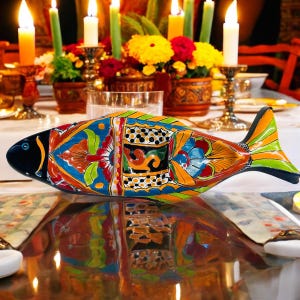 May include: A colorful, ceramic fish-shaped serving dish. The dish features a vibrant design with blue, orange, green, and yellow patterns. The fish has a black head and tail, and is resting on a reflective surface, with candles and flowers in the background.