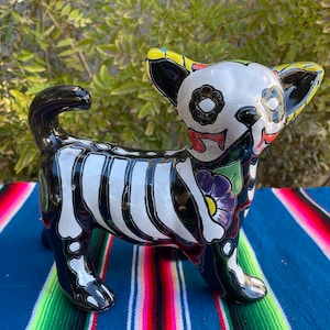 May include: A ceramic figurine of a chihuahua, painted in a Day of the Dead style. The dog is black and white with a skeleton design and colorful floral accents.